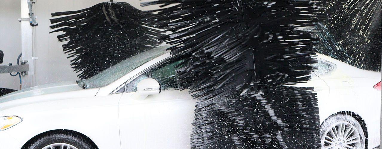 A white car being cleaned by large, rotating black brushes in an automatic car wash.